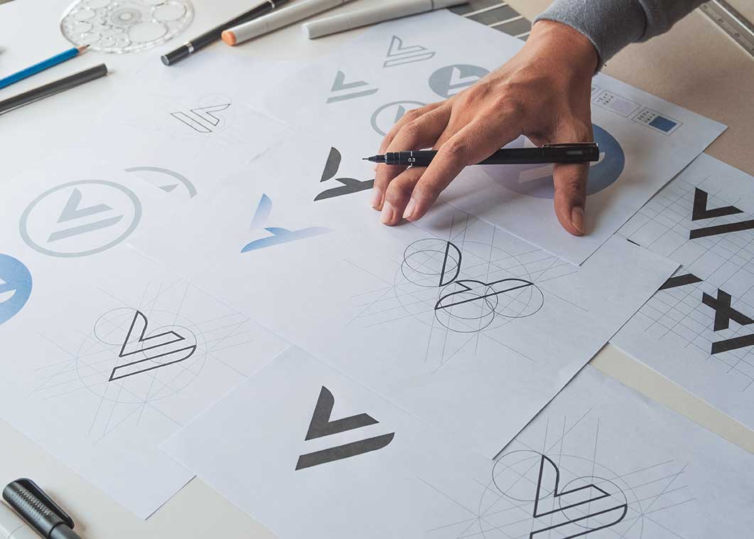 main d'un directeur artistique tenant un crayon au-dessus de plusieurs croquis de logos